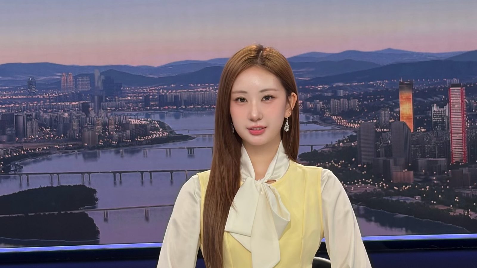 노란 조끼와 흰 리본 블라우스 차림으로 한강 야경 배경 앞에 선 전유진