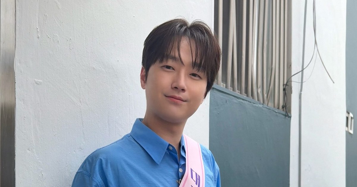 파란 셔츠에 어깨띠를 두르고 흰 벽 앞에서 미소 짓는 이찬원