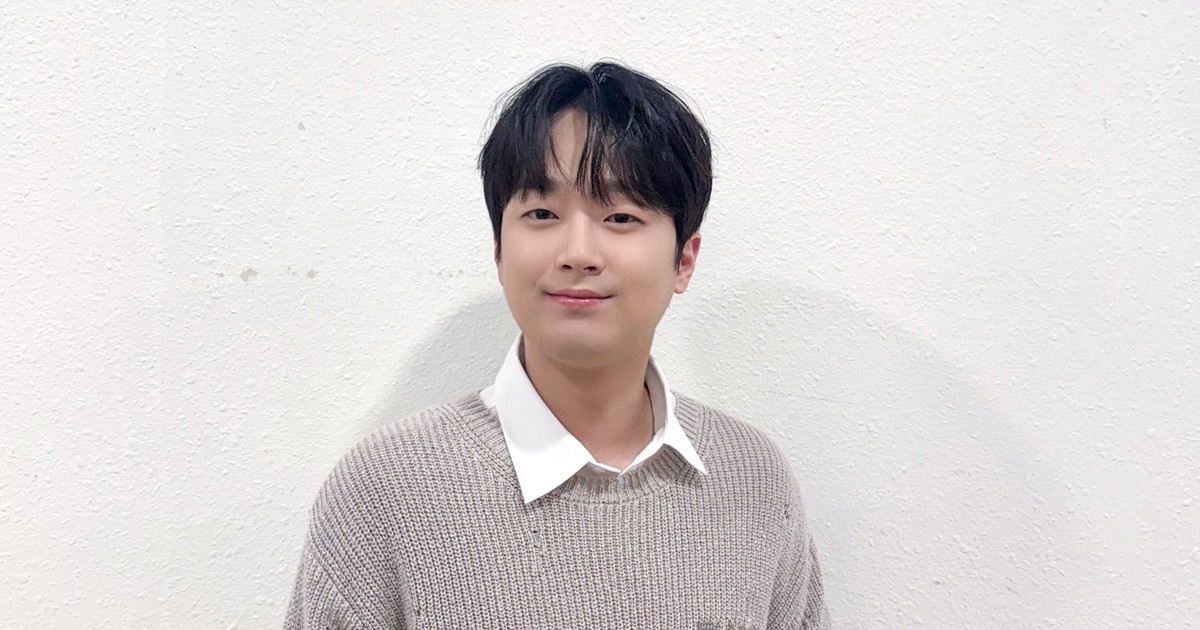 흰 벽 앞에서 베이지색 니트와 흰 셔츠를 입고 정면을 보며 미소 짓는 이찬원
