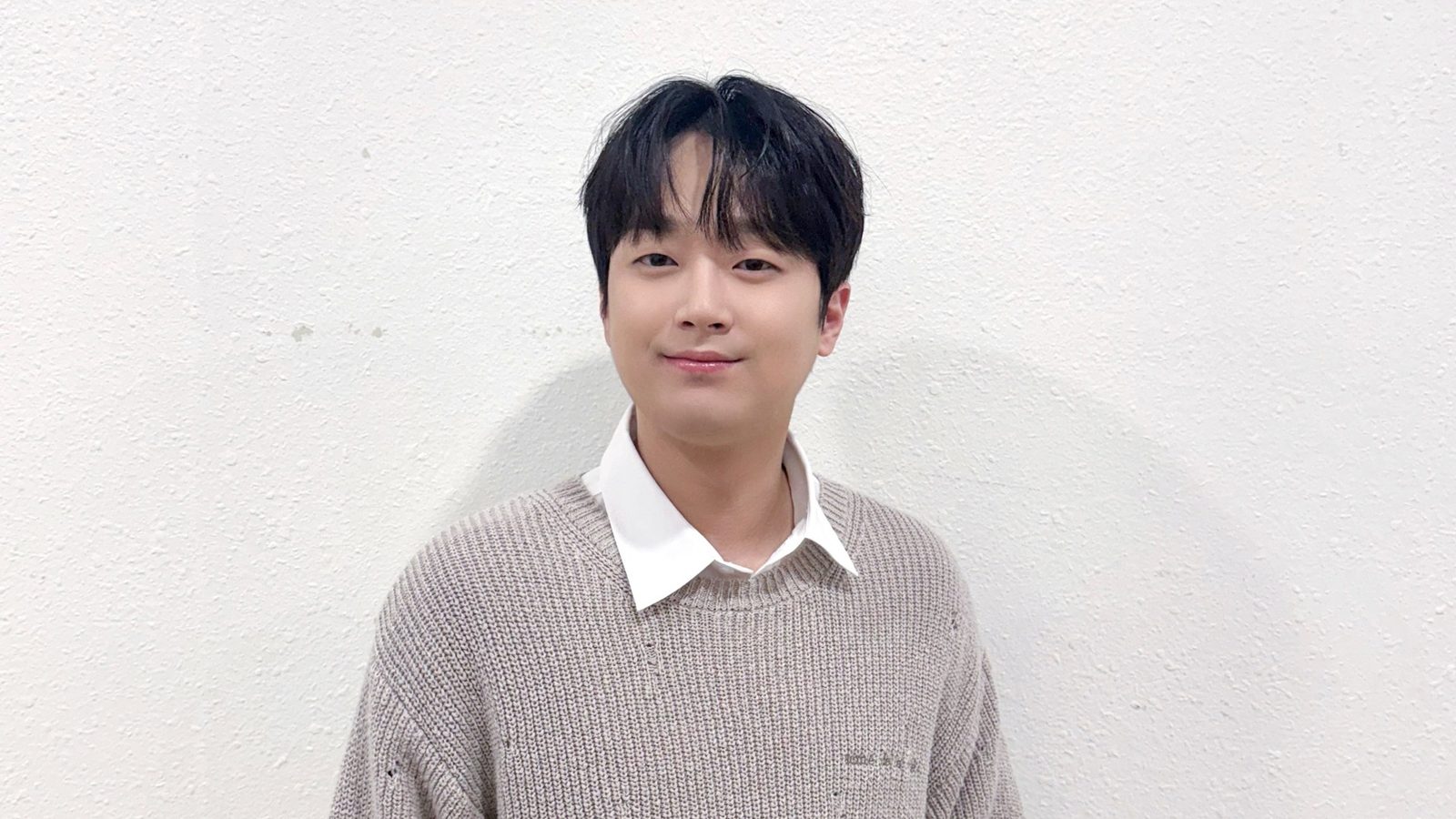 흰 벽 앞에서 베이지색 니트와 흰 셔츠를 입고 정면을 보며 미소 짓는 이찬원